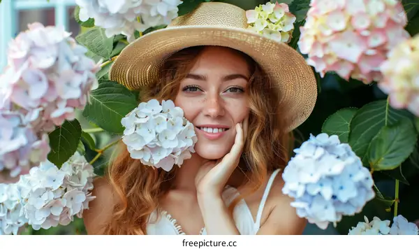 A beautiful young woman with freckles on her face is smiling. She is wearing a straw hat and is surrounded by colorful flowers.
