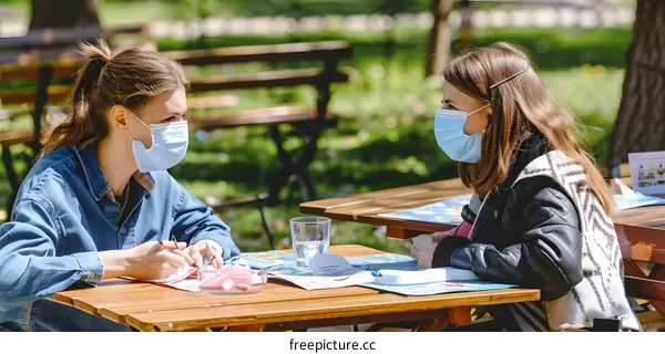 Two Young Women Wearing Face Masks Sit At An Outdoor Table