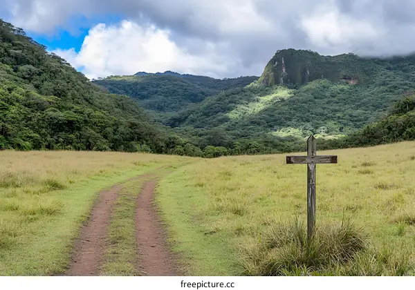 Dirt Road Leading Up to Wooden Cross in Front of Green Mountains