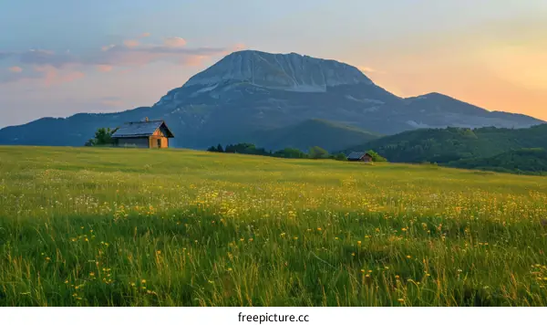 A serene mountain hut among a lush green valley with a mountain backdrop and a glorious sunset