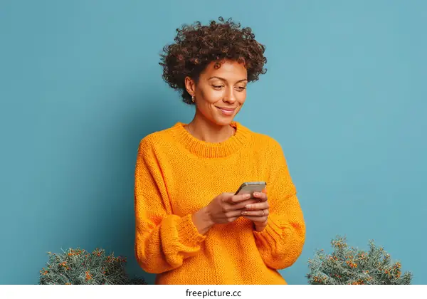 Happy young woman in orange sweater using smartphone against blue background