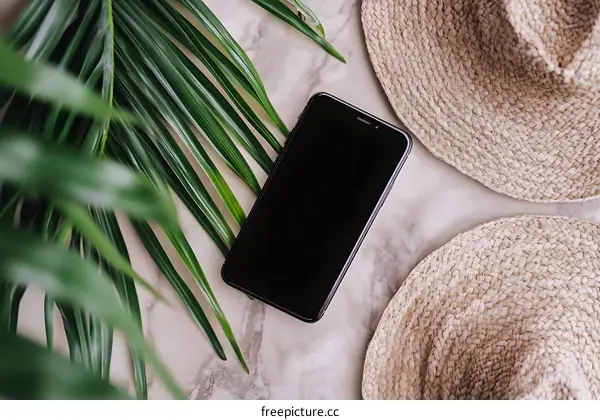 Black Smartphone on Marble Surface with Palm Leaves and Straw Hats