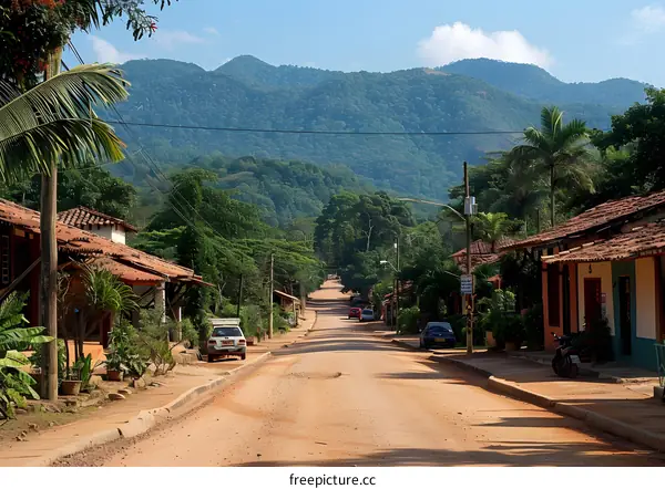 A long village road with cars parked on either side and a lush green mountain in the background
