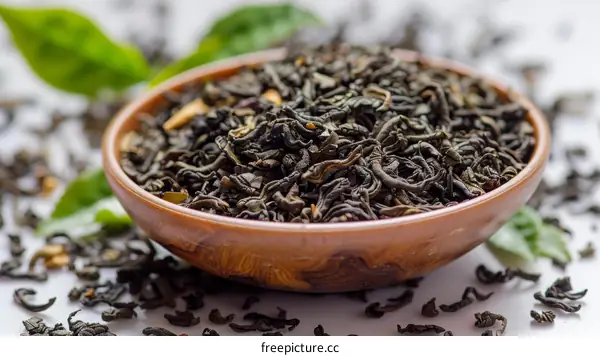 Black tea leaves in a wooden bowl with scattered tea leaves on the white surface