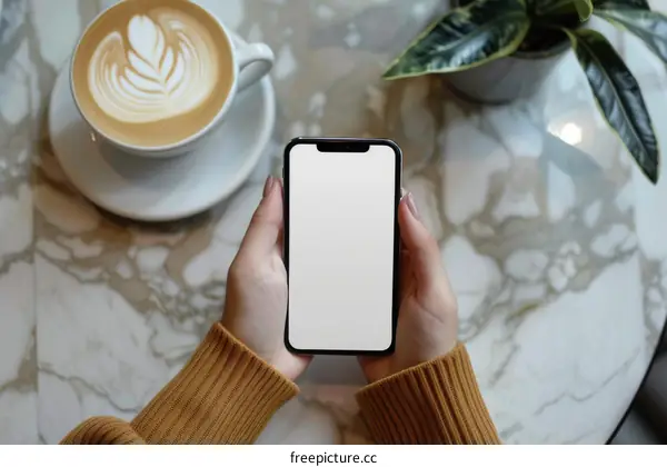 A person holding a smartphone with a cup of coffee and a plant on the table