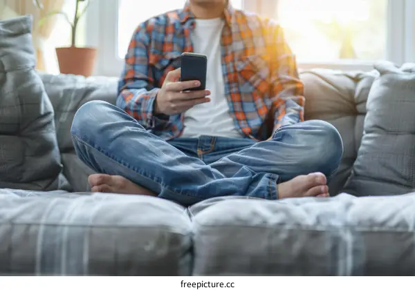A man in casual clothes is sitting on a couch and using a smartphone