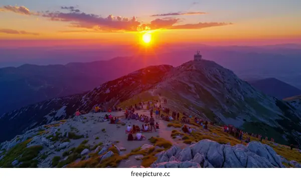 Crowds of people gather on a mountaintop to watch the sunset over a mountain range.