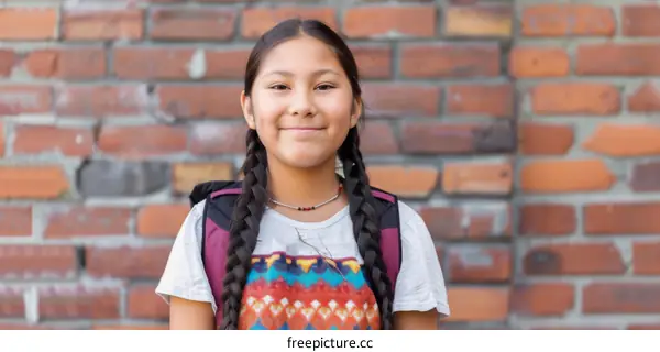 portrait of a smiling young native american girl with long black hair in braids wearing a colorful beaded necklace and a backpack standing in front of a brick wall