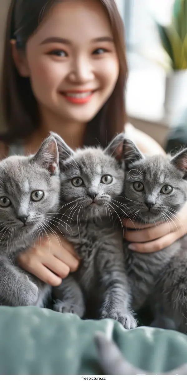 A young woman is holding three gray kittens in her arms and smiling at the camera.