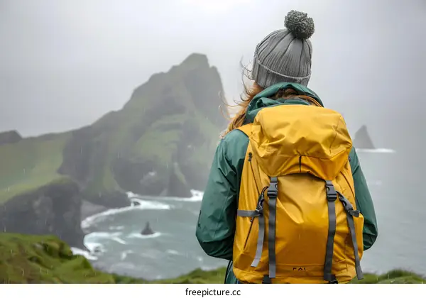 Woman in a Green Jacket and a Grey Beanie Standing on a Cliff Overlooking the Ocean on a Cloudy Day