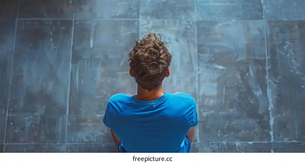 A young man sits on the floor with his back to the camera