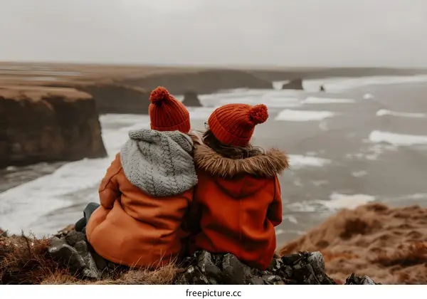 Two Women in Red Hats Sitting on a Cliff Overlooking the Ocean
