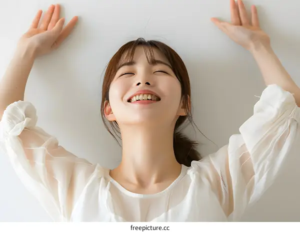 A young woman in a white dress is smiling with her eyes closed and her arms in the air
