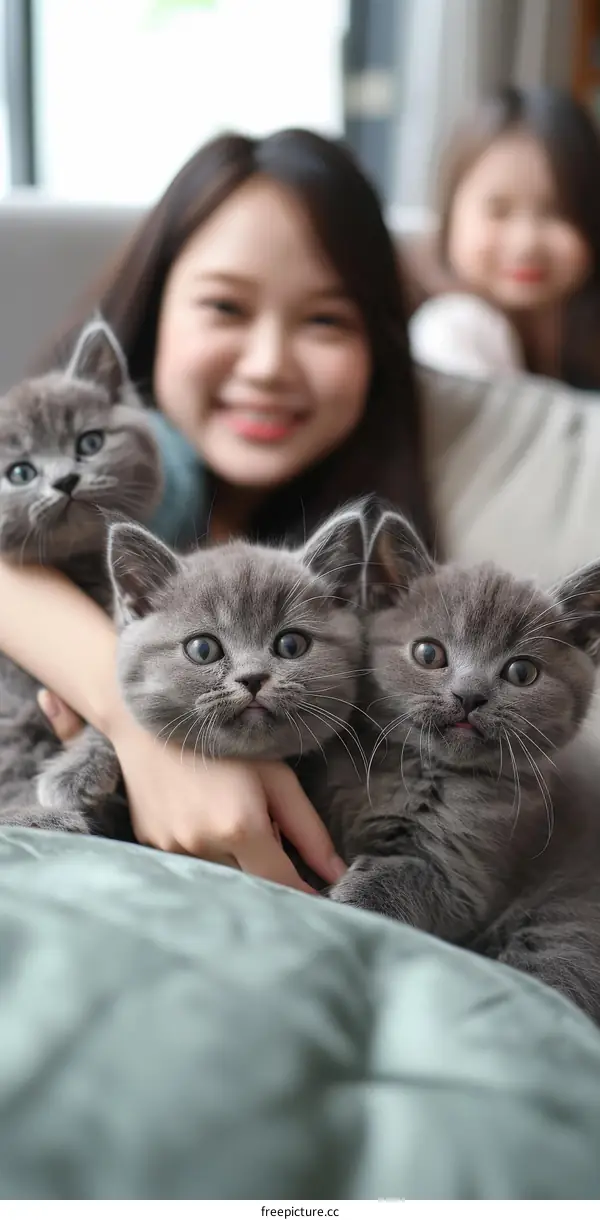 A young woman is holding three gray kittens in her arms and smiling at the camera.