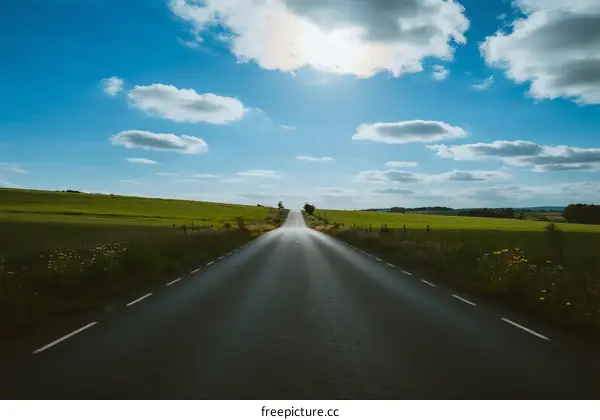 A long asphalt road stretching through green fields under a blue sky with clouds