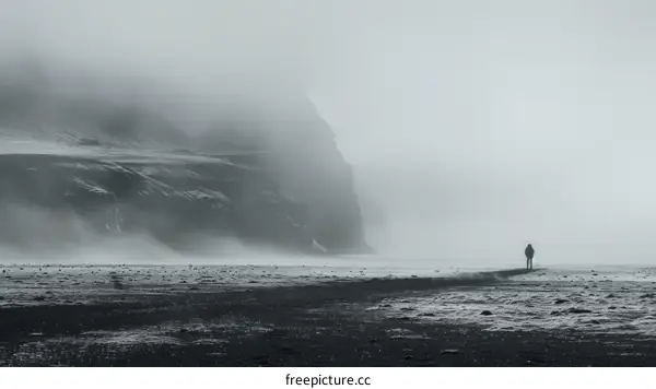 Black and white photo of a person standing alone on a beach with a large rock formation in the distance
