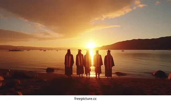 Five people in graduation gowns standing on a beach at sunset